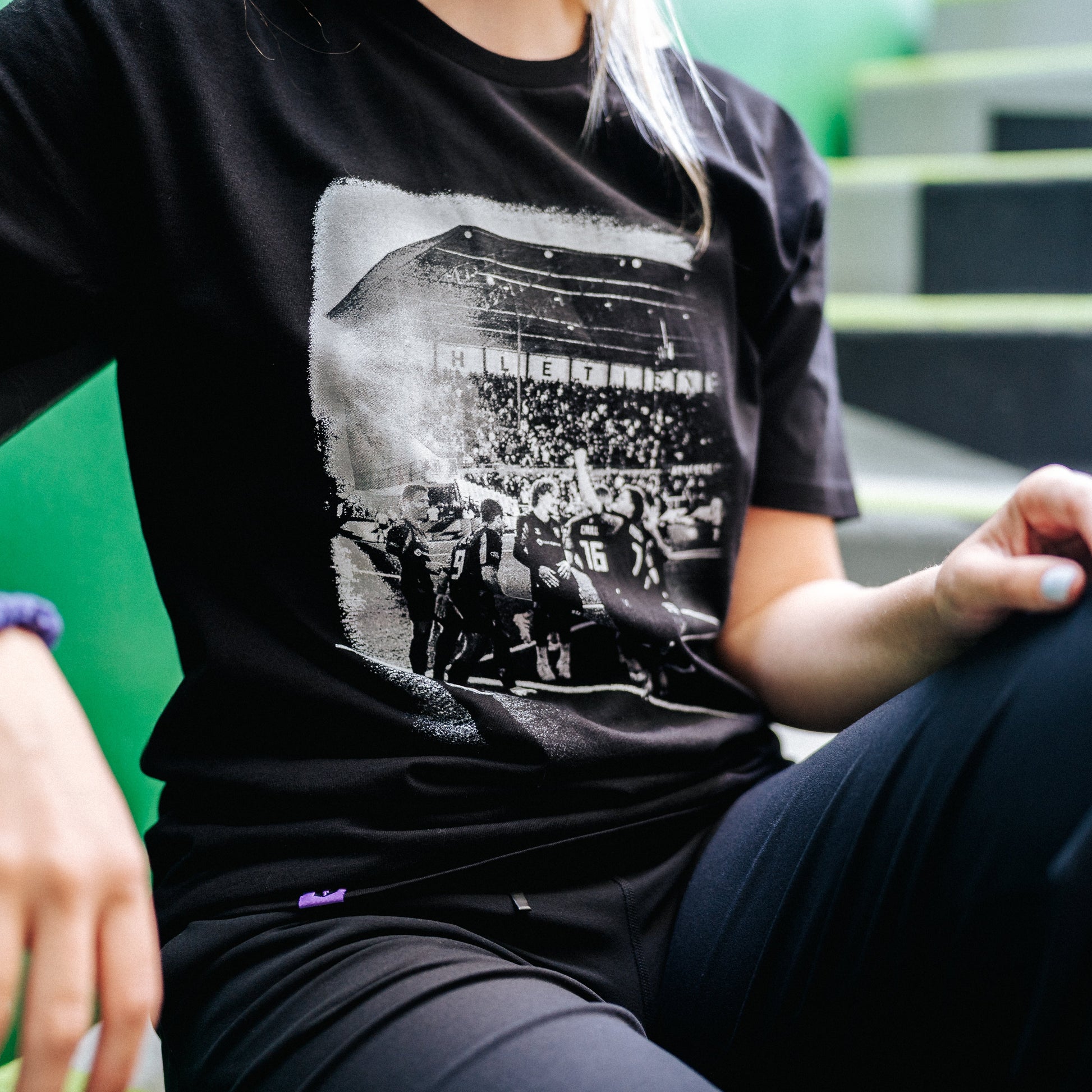 A person wearing a black graphic t-shirt with a black and white photo print of a crowd scene, seated against a bright green wall.