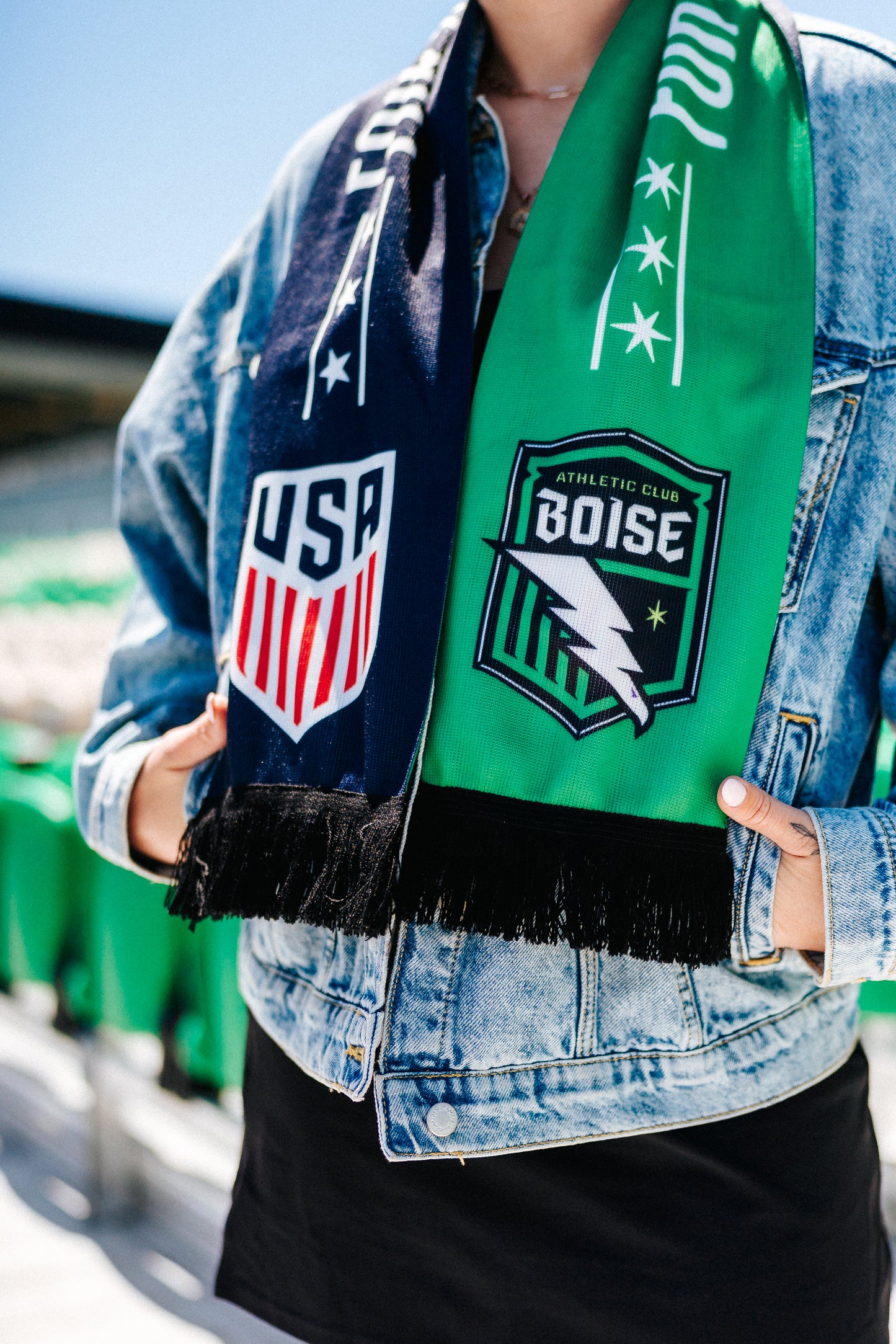 Person wearing a denim jacket with USA and Boise athletic club logos, holding a green scarf.